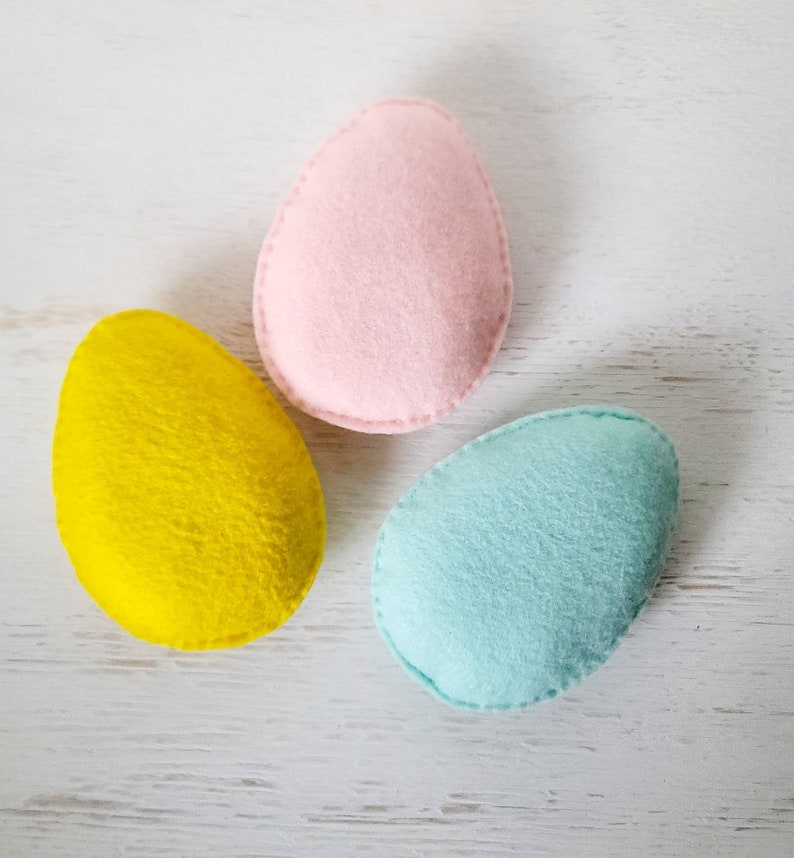 May include: Three felt Easter eggs in pink, yellow, and light blue. The eggs are arranged in a triangle shape on a white wooden surface.