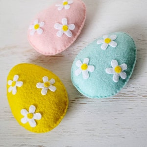 May include: Three felt Easter eggs, one pink, one light blue, and one yellow, each decorated with white felt flowers with yellow centers.