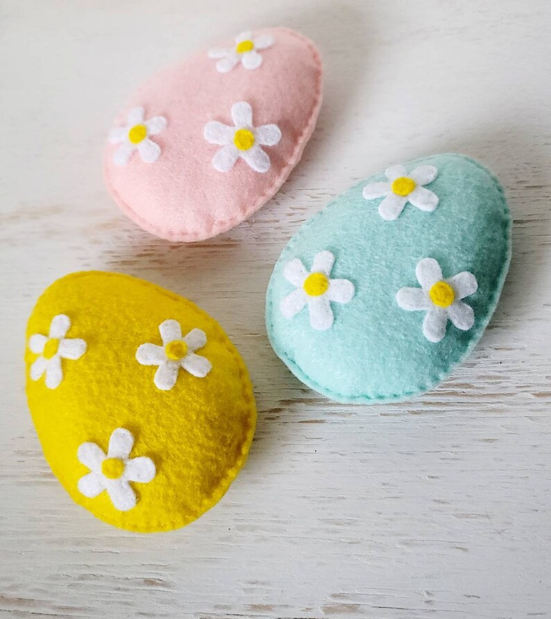 May include: Three felt Easter eggs, one pink, one light blue, and one yellow, each decorated with white felt flowers with yellow centers.