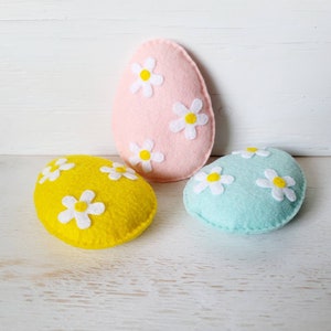 May include: Three felt Easter eggs, one pink, one yellow, and one light blue, each decorated with white felt flowers.