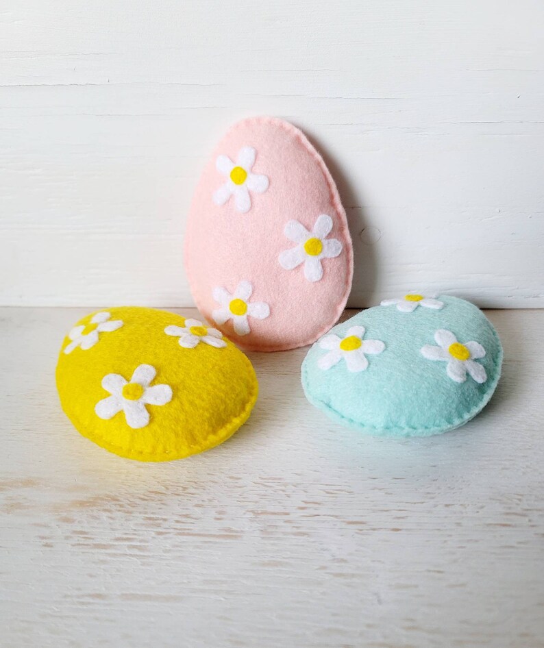 May include: Three felt Easter eggs, one pink, one yellow, and one light blue, each decorated with white felt flowers.
