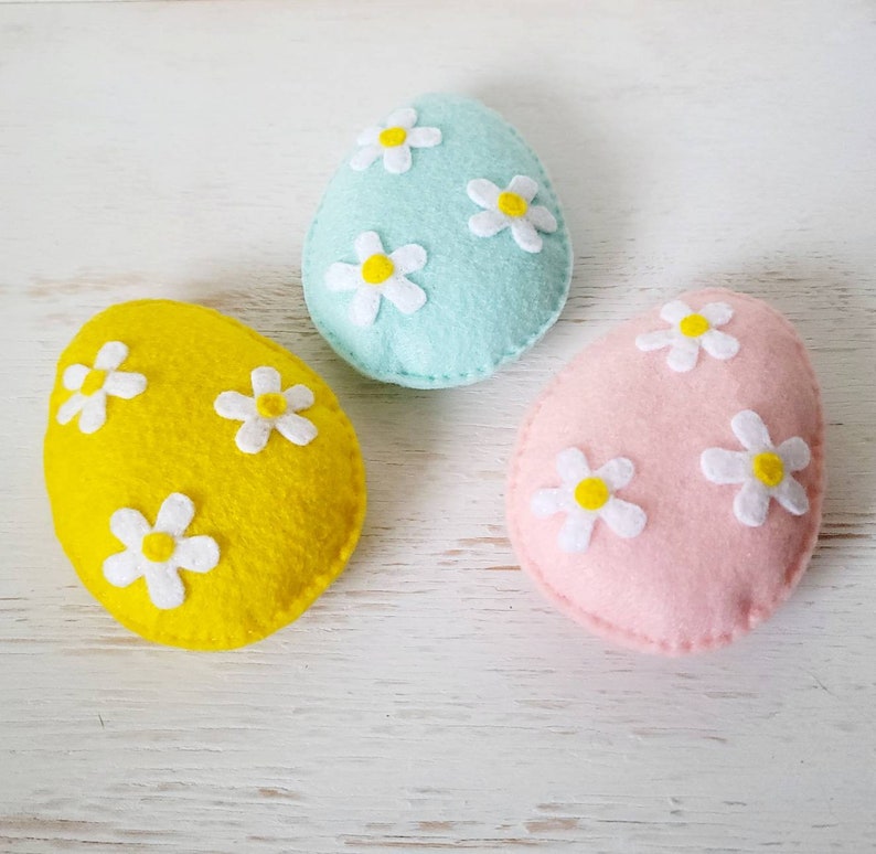 May include: Three felt Easter eggs, one yellow, one light blue, and one pink, each decorated with white felt flowers.