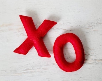 May include: Red felt letters 'X' and 'O' on a white wooden surface. The 'X' is slightly angled, and the 'O' is a perfect circle. The letters are likely handmade and could be used for Valentine's Day or other romantic occasions.