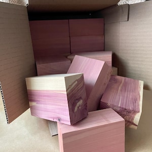 May include: A cardboard box filled with several rectangular cedar wood blocks. The blocks are a reddish-pink color with visible wood grain. Some blocks have a lighter, natural wood color on one side.