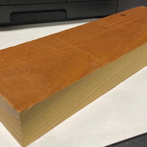 May include: A rectangular block of wood with a reddish-brown top surface and a lighter, yellowish-brown side. The wood grain is visible on the top. The block appears to be approximately 10 inches long.