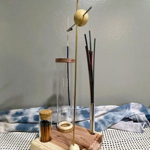 May include: A wooden incense holder with two glass jars, one containing matches and the other a long incense stick. The holder has a brass rod with three wooden spheres and a metal rod with a bunch of incense sticks.