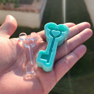 Op de afbeelding: Een turquoise blauwe siliconen mal in de vorm van een sleutel met een hart in het midden en Mickey Mouse oren aan de bovenkant. Naast de mal ligt een transparante plastic mal met hetzelfde ontwerp.