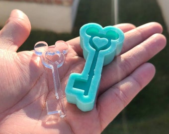 May include: A turquoise blue silicone mold shaped like a key with a heart in the middle and Mickey Mouse ears at the top. A clear plastic mold of the same design is next to it.