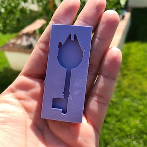 Pode incluir: Um molde de silicone de cor lavanda em forma de chave. O design da chave apresenta um topo detalhado que se assemelha a um edifício. O molde retangular é segurado em uma mão, com um fundo verde desfocado.