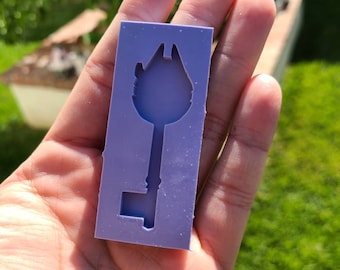 May include: A lavender-colored silicone mold in the shape of a key. The key design features a detailed top resembling a building. The mold is rectangular and held in a hand, with a blurred green background.