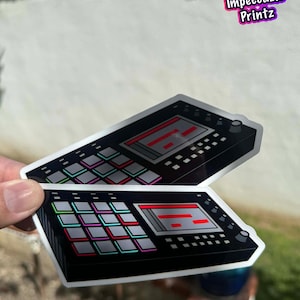 Puede incluir: Dos pegatinas con un diseño de dispositivo de producción de música electrónica en negro y gris. Las pegatinas tienen una combinación de colores rojo y verde y están etiquetadas como "Pack de 2". Las pegatinas se sostienen contra un fondo borroso.