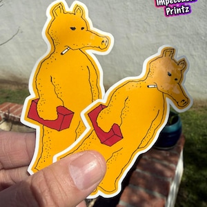 Puede incluir: Dos pegatinas amarillas con un personaje de dibujos animados parecido a un oso sosteniendo un bloque rojo y un cigarrillo. Las pegatinas tienen un borde blanco y están etiquetadas como "Pack of 2" e "Impeccable Printz".