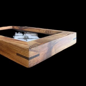 Könnte beinhalten: Ein Holzbilderrahmen mit dunklem Innenraum. Der Rahmen besteht aus hellbraunem Holz mit sichtbarer Maserung und dunklen grauen Eckakzenten. Der Rahmen ist vor einem schwarzen Hintergrund platziert.