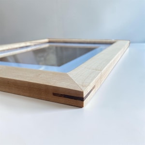 May include: A light-colored wooden picture frame with a glass insert. The frame has a simple, modern design with a natural wood grain finish. The corners are joined with a dark brown accent. The frame is angled, showing its construction.
