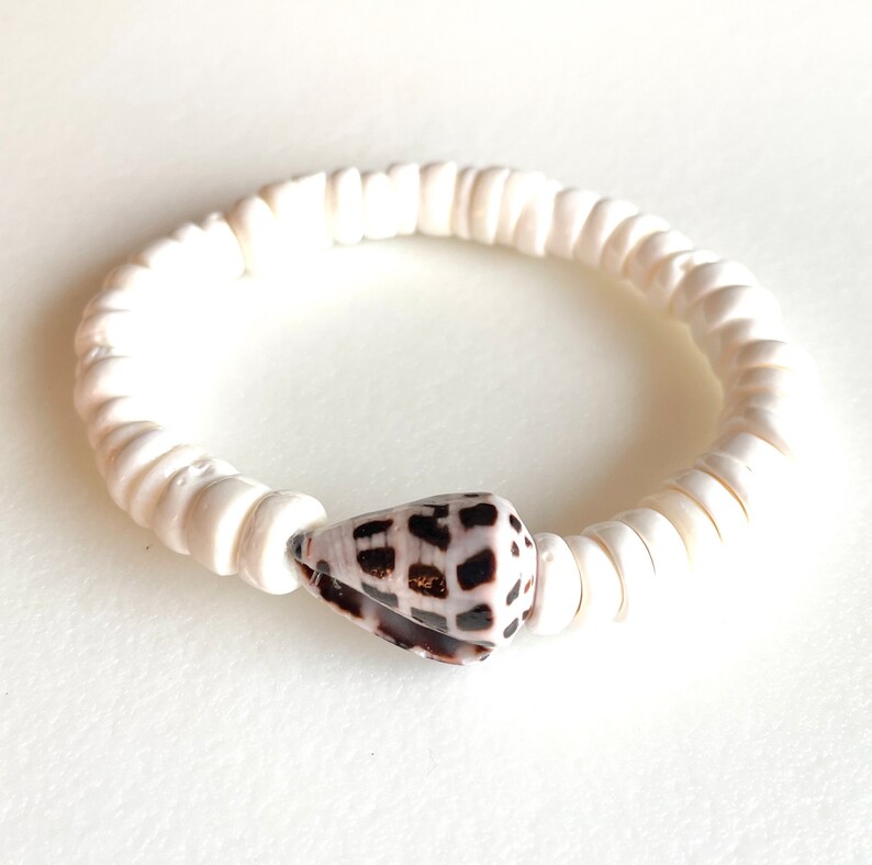 K&ouml;nnte beinhalten: Armband aus wei&szlig;en Muschelkernperlen mit einem braunen und wei&szlig;en Muschel-Anh&auml;nger.