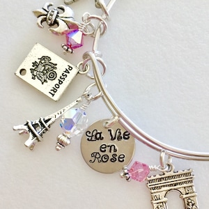 Op de afbeelding: Een zilveren armband met verschillende bedels, waaronder de Eiffeltoren, een paspoort, een fleur-de-lis, een kristallen kraal en een bedel met de tekst "La Vie en Rose".