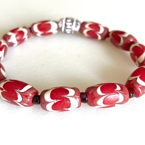 May include: A red and white patterned bracelet with a silver bead charm. The bracelet is made of oval-shaped beads with a white swirl design on a red background.
