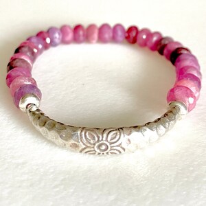 Könnte beinhalten: Ein Armband aus rosafarbenen und violetten Edelsteinen mit einem silbernen Blumenschloss. Das Armband besteht aus kleinen, runden Perlen und hat ein gehämmertes Silberband.