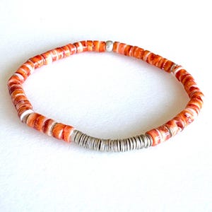 May include: A beaded bracelet featuring orange and white shell beads strung together with silver-colored metal spacers. The bracelet is a simple, circular design, perfect for everyday wear. The beads are approximately 0.25 inches in diameter.
