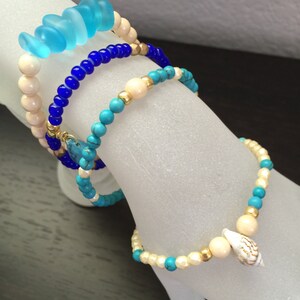May include: Four bracelets stacked on a white surface. The top bracelet is made of light blue glass beads. The second bracelet is made of dark blue beads. The third bracelet is made of turquoise and white beads. The bottom bracelet is made of white and gold beads with a small seashell charm.