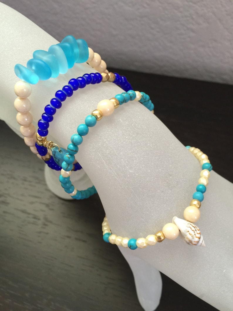 May include: Four bracelets stacked on a white surface. The top bracelet is made of light blue glass beads. The second bracelet is made of dark blue beads. The third bracelet is made of turquoise and white beads. The bottom bracelet is made of white and gold beads with a small seashell charm.