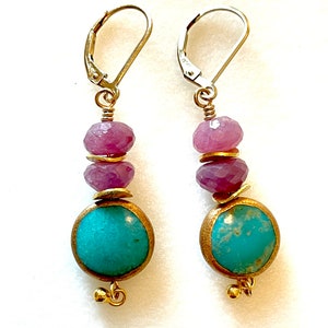 May include: A pair of dangle earrings with a turquoise and pink gemstone design. The earrings feature a round turquoise stone with gold accents and two pink gemstones above it. The earrings are attached to silver leverback closures.