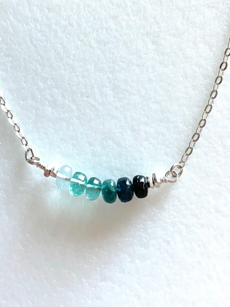 May include: A silver chain necklace with a graduated row of blue gemstones. The stones range in colour from light blue to dark blue.