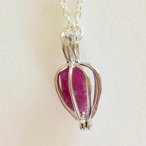 Könnte beinhalten: Eine silberne Halskette mit einem Käfig-Anhänger, der einen rosa Kristall enthält.