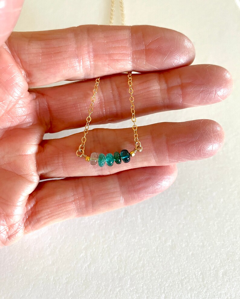 May include: A gold chain necklace with a row of graduated green gemstones. The necklace is delicate and features a subtle ombre effect.