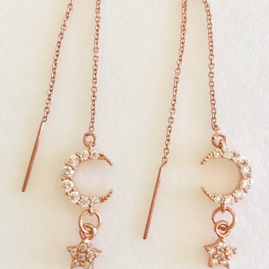 Puede incluir: Un par de pendientes colgantes de oro rosa con un dije de media luna y estrella. La luna y la estrella están incrustadas con diamantes de imitación transparentes.