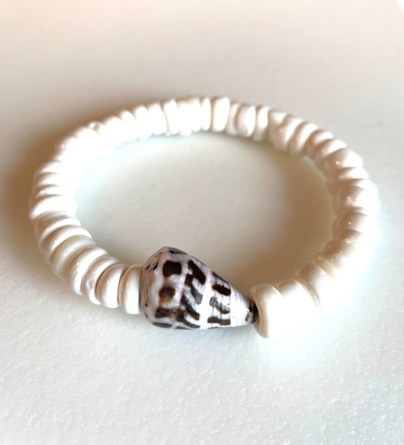 K&ouml;nnte beinhalten: Armband aus wei&szlig;en Muschelperlen mit einem braunen und wei&szlig;en Muschel-Anh&auml;nger.