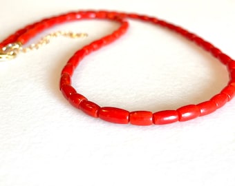 May include: A bright red necklace featuring small, cylindrical beads. The beads are strung together, forming a continuous loop with a gold-coloured clasp and extender chain. The necklace is displayed on a white surface.