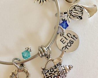 May include: A silver charm bracelet with various charms, including a pretzel, a beer stein, a flower, a hat, a horn, and a blue crystal. The bracelet also has a charm with the text "Ein Prosit".