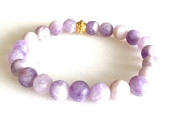 Puede incluir: Una pulsera de cuentas redondas de color lavanda. Una pequeña cuenta dorada se coloca en el centro. La pulsera se muestra sobre un fondo blanco, resaltando los colores y texturas de la joya.