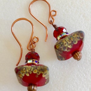 May include: A pair of red and gold dangle earrings with copper wire and hooks. The earrings feature a red glass bead with gold accents and a smaller red crystal bead.