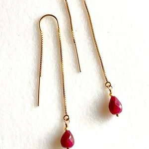 Op de afbeelding: Een paar gouden kettingoorbellen met een traanvormige robijn-edelsteen aan het einde van elke ketting.