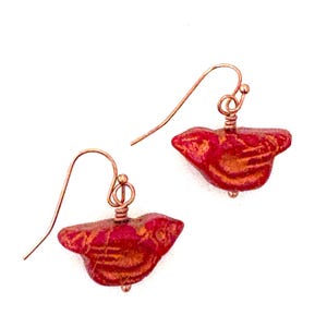 May include: A pair of red and gold-toned earrings with a bird-like shape. The earrings have copper-colored ear wires and are set against a white background. The earrings are a unique jewelry item.