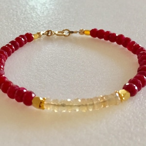 May include: A red and gold beaded bracelet with a gold clasp. The bracelet is made with small, faceted red beads and a row of smaller, clear beads.