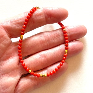 Pode incluir: Uma pulseira delicada com pequenas contas laranja vibrantes intercaladas com contas douradas geom&eacute;tricas. A pulseira &eacute; enfiada em uma corrente fina e foi projetada para ser usada ao redor do pulso. A joia &eacute; exibida em um fundo neutro.