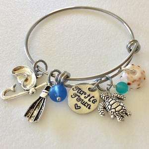 Può includere: Un braccialetto con ciondoli in argento con una varietà di ciondoli, tra cui una conchiglia, una tartaruga, una perla di vetro blu, una chiave, una pinna e un ciondolo che dice "Tur'tle Town".