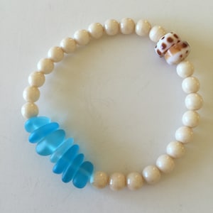 May include: A white bead bracelet with a blue glass accent and a brown and white seashell charm.