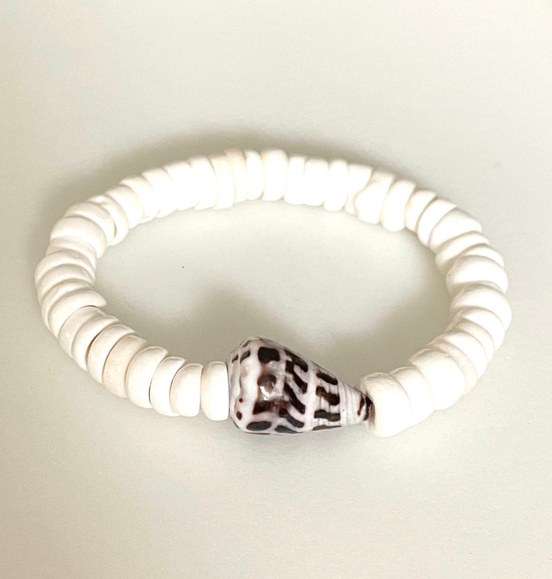K&ouml;nnte beinhalten: Armband aus wei&szlig;en Muschelkernperlen mit einem einzigen braunen und wei&szlig;en Muschel-Anh&auml;nger.