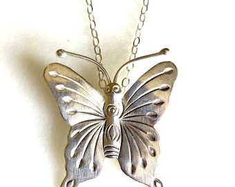 May include: A silver butterfly pendant necklace with a detailed design. The butterfly has a textured surface and intricate details on its wings.