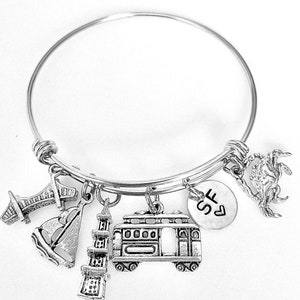 Puede incluir: Pulsera de plata con varios encantos, incluyendo un tranvía, un puente, un faro, un velero, un cangrejo y un encanto con las letras "SF" y un corazón.