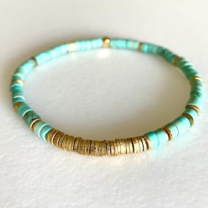 May include: A turquoise and gold beaded bracelet. The bracelet is made of small, round beads that are strung together on a thin cord. The beads are a mix of turquoise and gold, and the bracelet has a simple, elegant design.