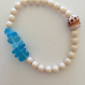 May include: A white beaded bracelet with a blue glass charm and a brown seashell charm.