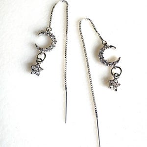 Puede incluir: Un par de pendientes colgantes plateados. Cada pendiente presenta una luna creciente y un charm de estrella de cinco puntas, ambos incrustados con pequeños cristales transparentes. Los pendientes cuelgan de delicadas cadenas.