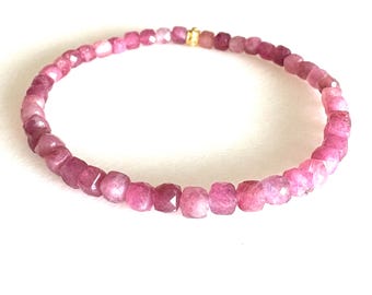 May include: A pink gemstone bracelet with faceted beads. The bracelet is made of small, square-cut pink gemstones strung together. A small gold-colored clasp secures the bracelet. The gemstones vary in shades of pink.