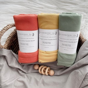 May include: Three rolled organic cotton swaddle blankets in a basket, in shades of orange, yellow, and green. Each blanket is labeled with the text "BLUMBERRY Co. BABY ESSENTIALS Organic Cotton Swaddle Blanket". A wooden baby rattle is in the foreground.