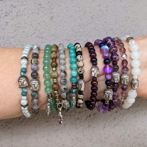 May include: A collection of beaded bracelets in various colors, including turquoise, green, purple, and white. Each bracelet features a different bead style and some include silver Buddha head charms. The bracelets are stacked on a wrist.